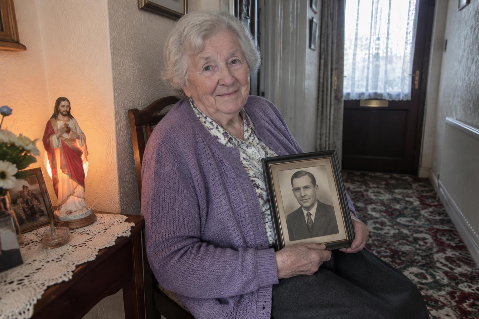 An older woman in her hallway holding a framed photo of her late husband